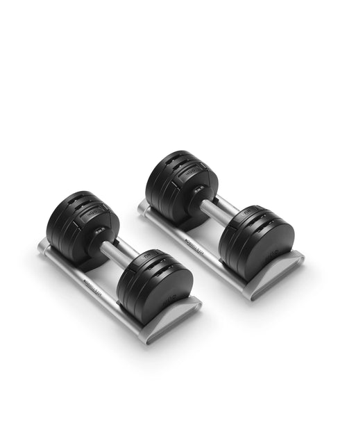 NÜOBELL S 224 adjustable dumbbell pair resting on silver storage trays against a white background, photographed from a three-quarter angle to display the layered black weight plates, brushed aluminum handles with visible branding, and the twist-dial selectors on both ends of each dumbbell.