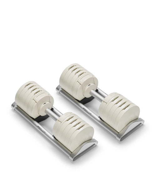 Side profile of two NÜOBELL S 240 adjustable dumbbells in cream finish positioned on silver trays, highlighting the machined aluminum handles, stacked weight plates with 2 kg increment markings, and compact footprint that replaces 20 pairs of fixed dumbbells.